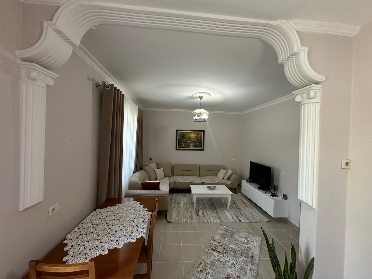A living area features a large, L-shaped sectional sofa and a coffee table, providing ample seating. Natural light enters through a window dressed with curtains. A dining table with wooden chairs and decorative plants are also present, enhancing the space's comfort.