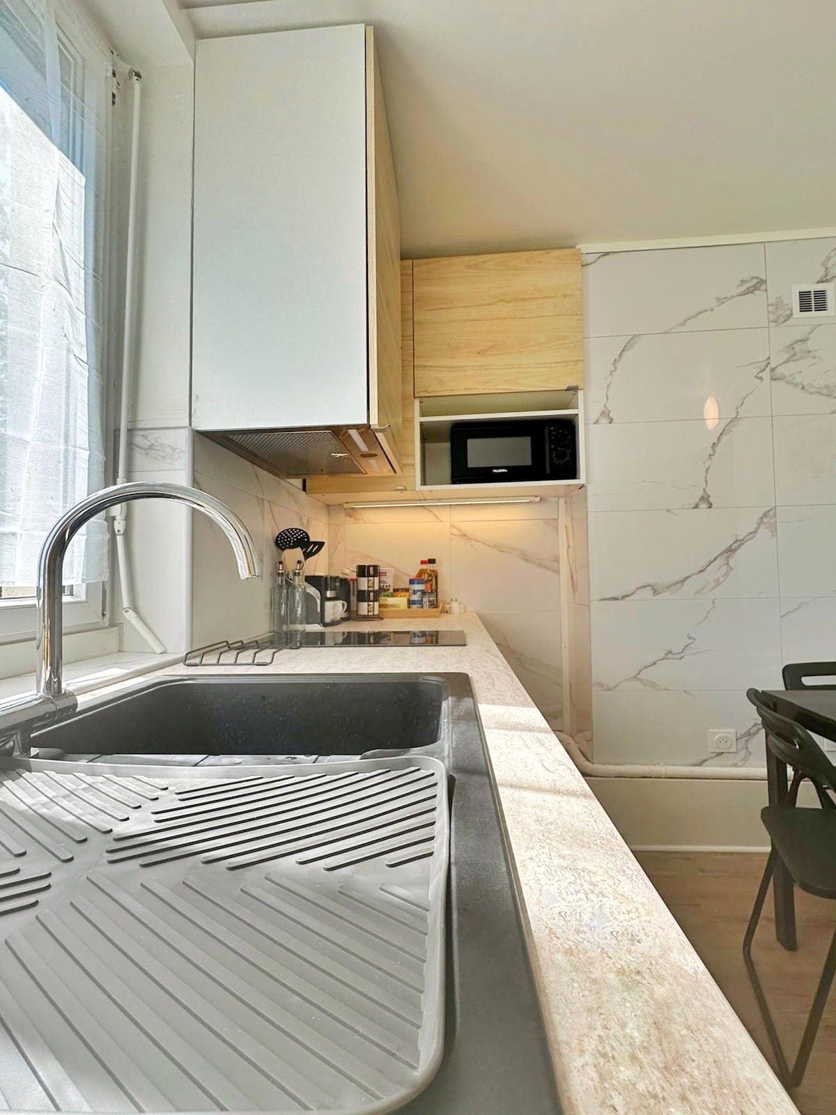 The kitchen features a modern sink with a sleek design. Cabinetry is visible above, providing storage space, while a microwave is placed on the countertop. Natural light filters through the window, illuminating the clean lines and marble backsplash of this functional cooking space.