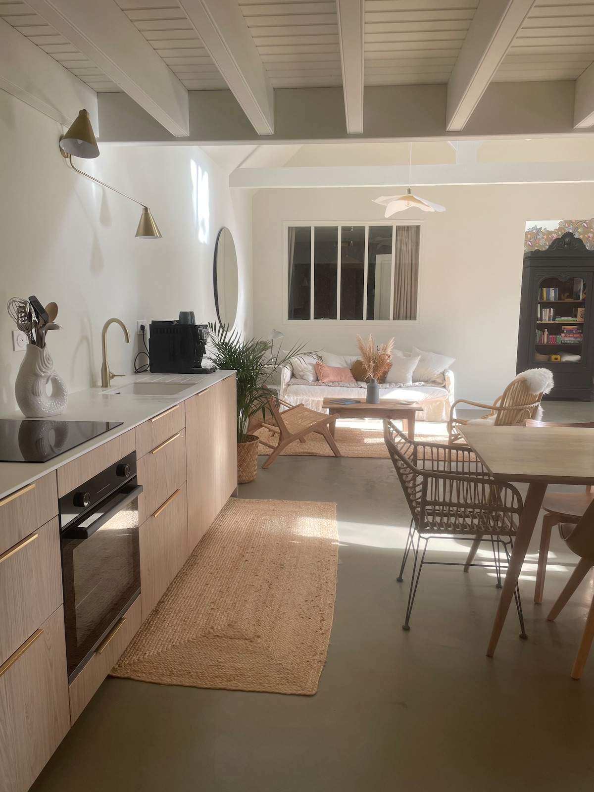 The kitchen area features modern appliances including an oven and a sleek sink. A cozy seating area with a light sofa and decorative elements is visible. Natural light brightens the space, creating a calm atmosphere, complemented by a dining table and woven chairs.