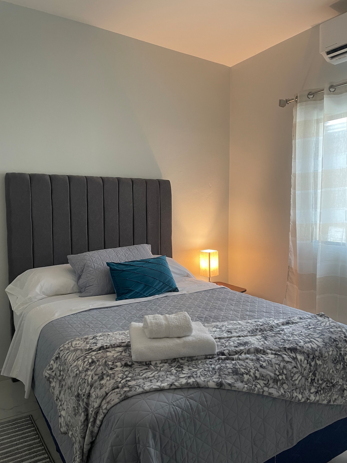 A cozy sleeping area features a gray tufted headboard above a spacious bed. The bedding is complemented by a decorative pillow in teal. Soft light is provided by a lamp on the bedside table, while a window dressed in sheer curtains allows natural light to filter in.