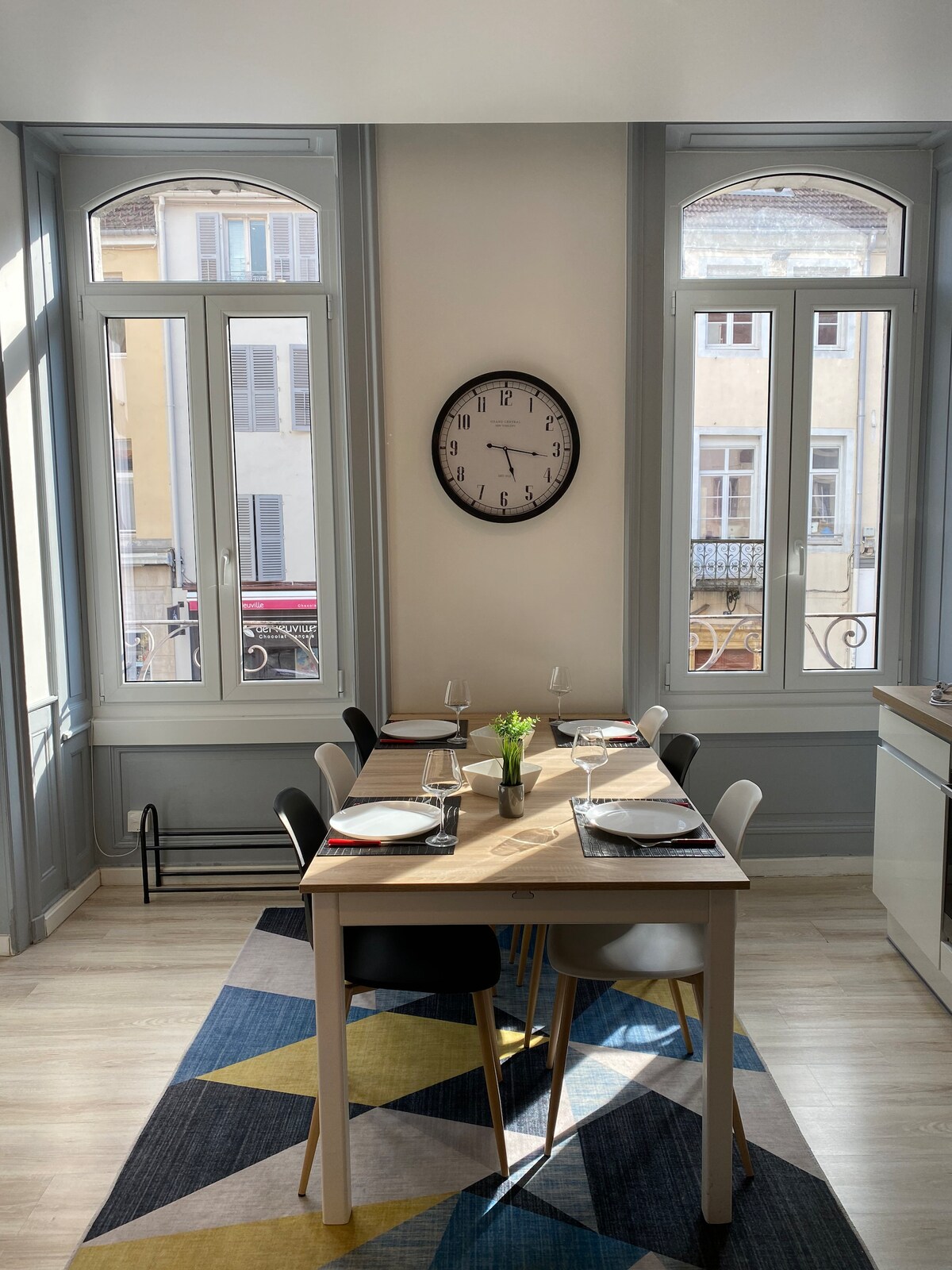 A dining area is illuminated by natural light, with large windows framed in light blue. A modern table is set for meals, complemented by a clock on the wall. Various chairs surround the table, with a patterned rug beneath adding a touch of colour.