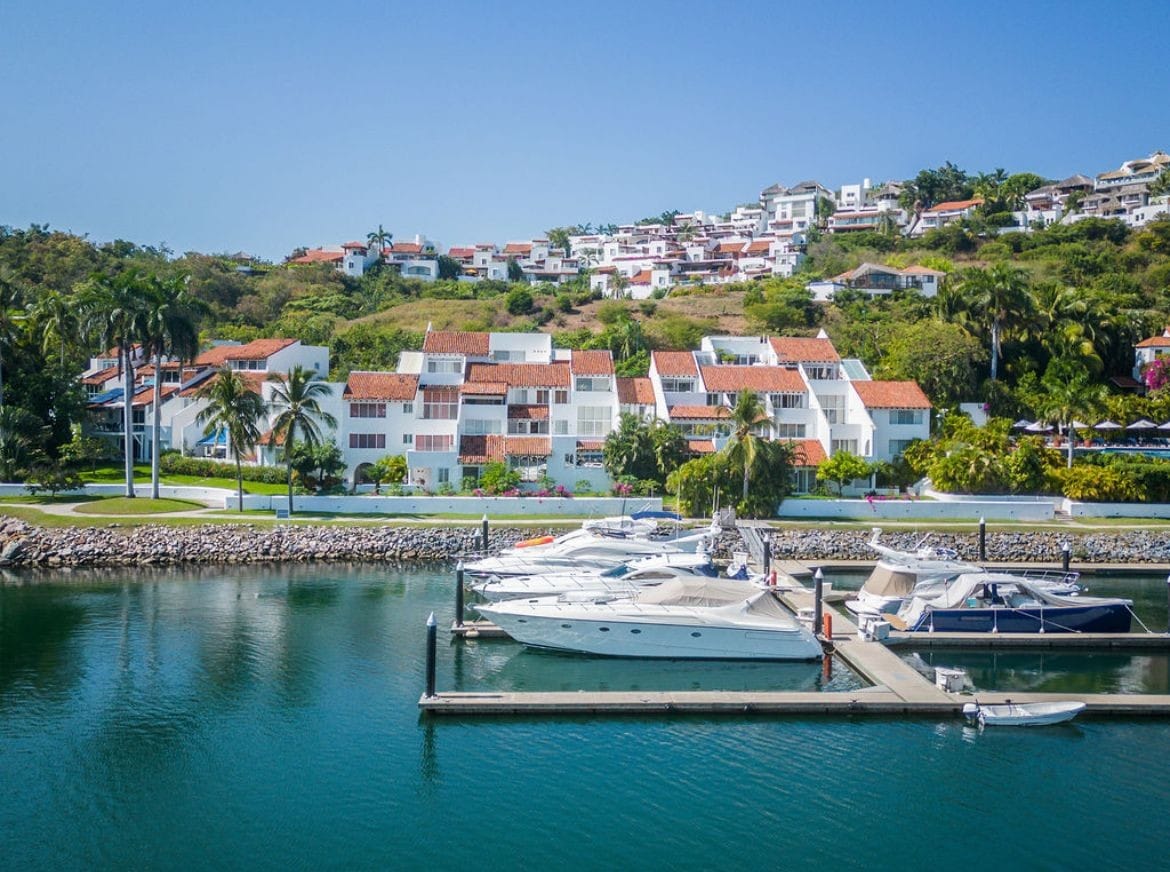 A scenic view showcases several boats docked at a marina, surrounded by calm, turquoise waters. Lush greenery and palm trees line the background, with a hillside of white buildings featuring red-tiled roofs visible above. A clear blue sky enhances the tranquil atmosphere.