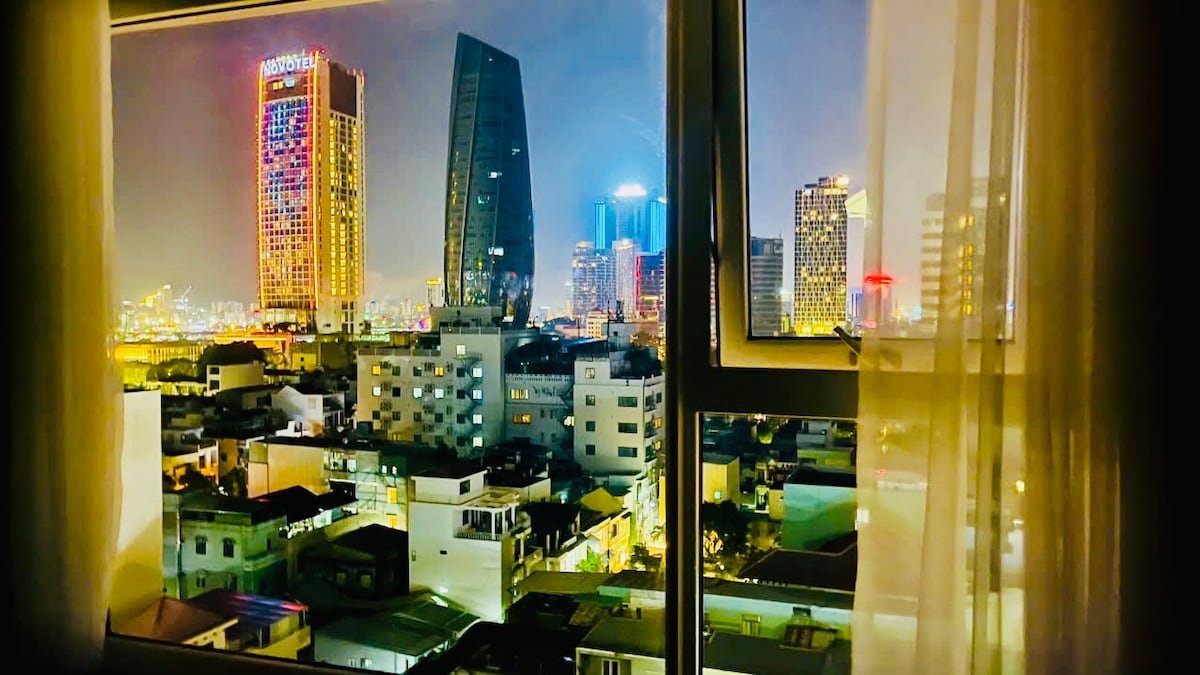 A city skyline is presented through a window, showcasing numerous buildings illuminated at night. Colorful lights from taller structures create a vibrant backdrop against the darker rooftops below. Sheer curtains softly frame the view, allowing glimpses of the bustling urban environment.