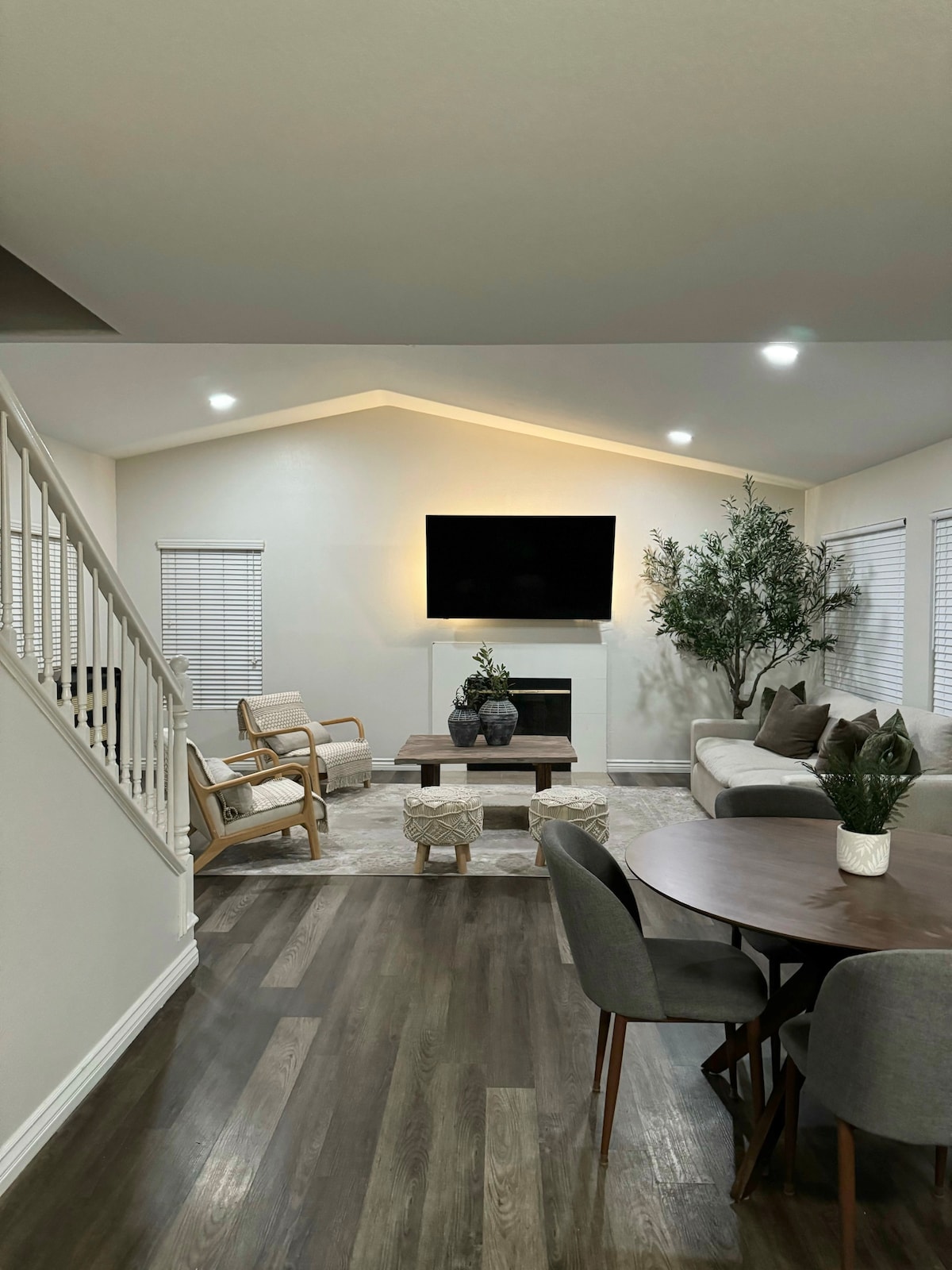 The living area features a light-filled space with modern furnishings. A large sectional sofa is positioned alongside a cozy fireplace. A round dining table with upholstered chairs adds functionality, while decorative plants and a coffee table complete the inviting atmosphere.