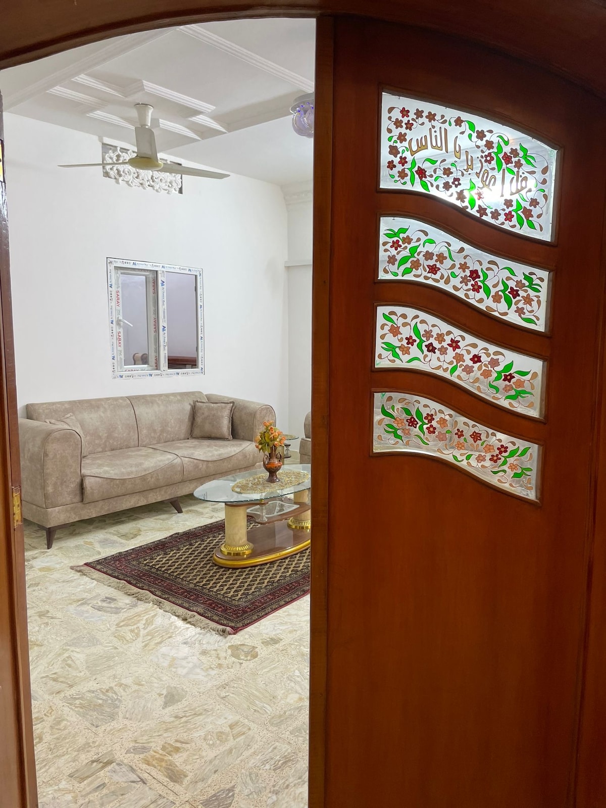A spacious living area is visible through a decorative wooden door featuring stained glass. A light-colored sofa is paired with a circular glass coffee table, while a colorful rug anchors the space. Natural light filters in, enhancing the room's openness.