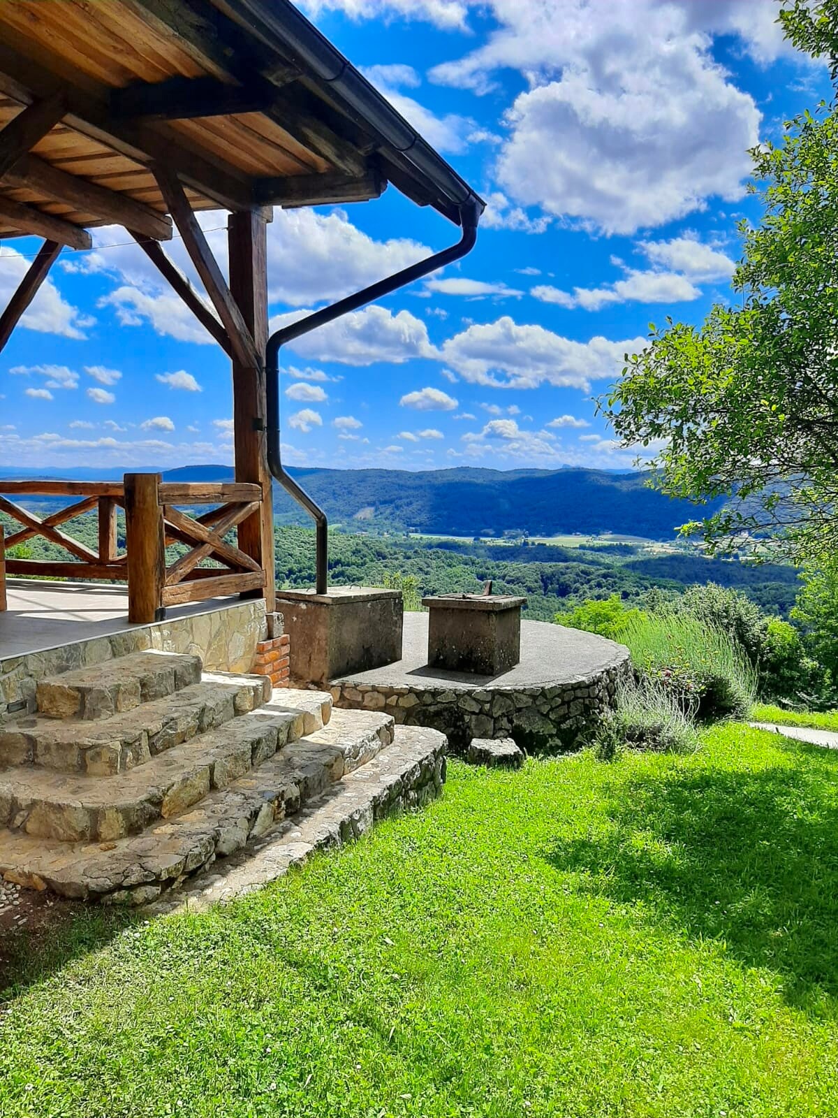 An outdoor area presents stone steps leading to a wooden gazebo, surrounded by lush greenery. Panoramic views of rolling hills and a bright blue sky with scattered clouds create a serene setting. Natural elements emphasize the peaceful atmosphere of the location.