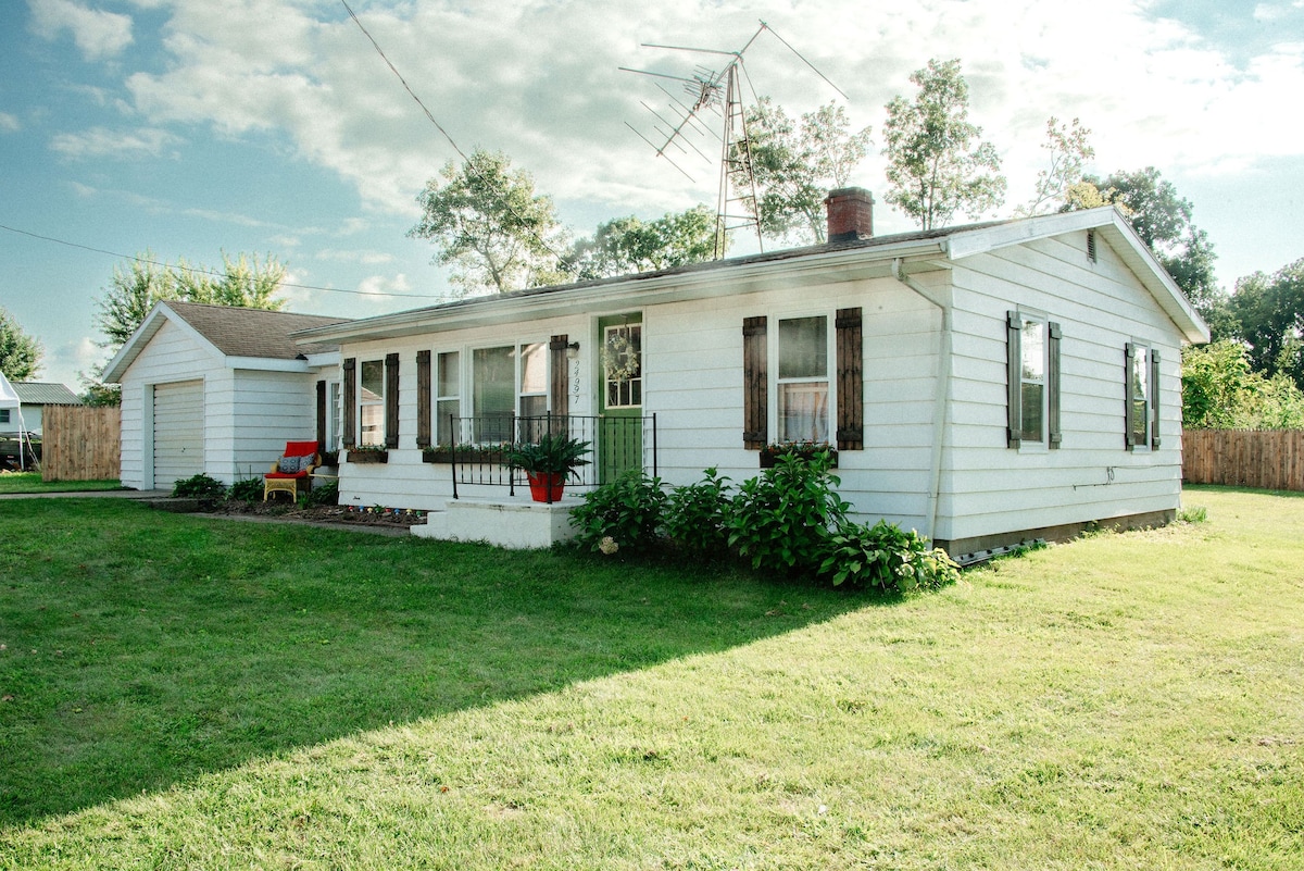 A quaint cottage is showcased with a well-maintained exterior, featuring white siding and decorative shutters. A welcoming porch with seating overlooks a grassy lawn, and a private driveway leads to a small garage. Surrounding trees provide a natural backdrop.