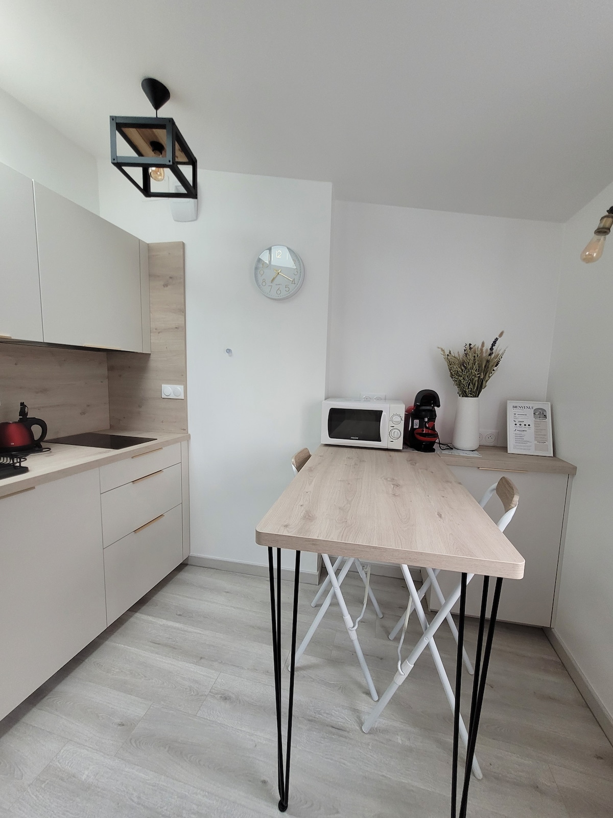 A compact kitchen area features minimalist cabinetry and a light wood countertop. A small table with three white chairs offers dining space. A microwave and a coffee machine are positioned on the countertop, and a wall clock adds functionality.