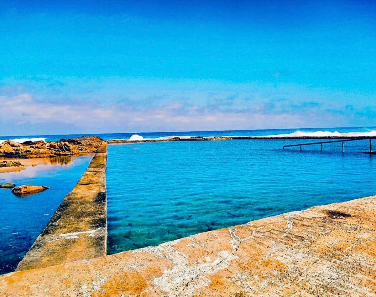 A clear blue pool is visible, bordered by a rugged stone walkway that leads to the ocean. Gentle waves can be seen in the distance, while a serene sky overhead is filled with soft, scattered clouds.