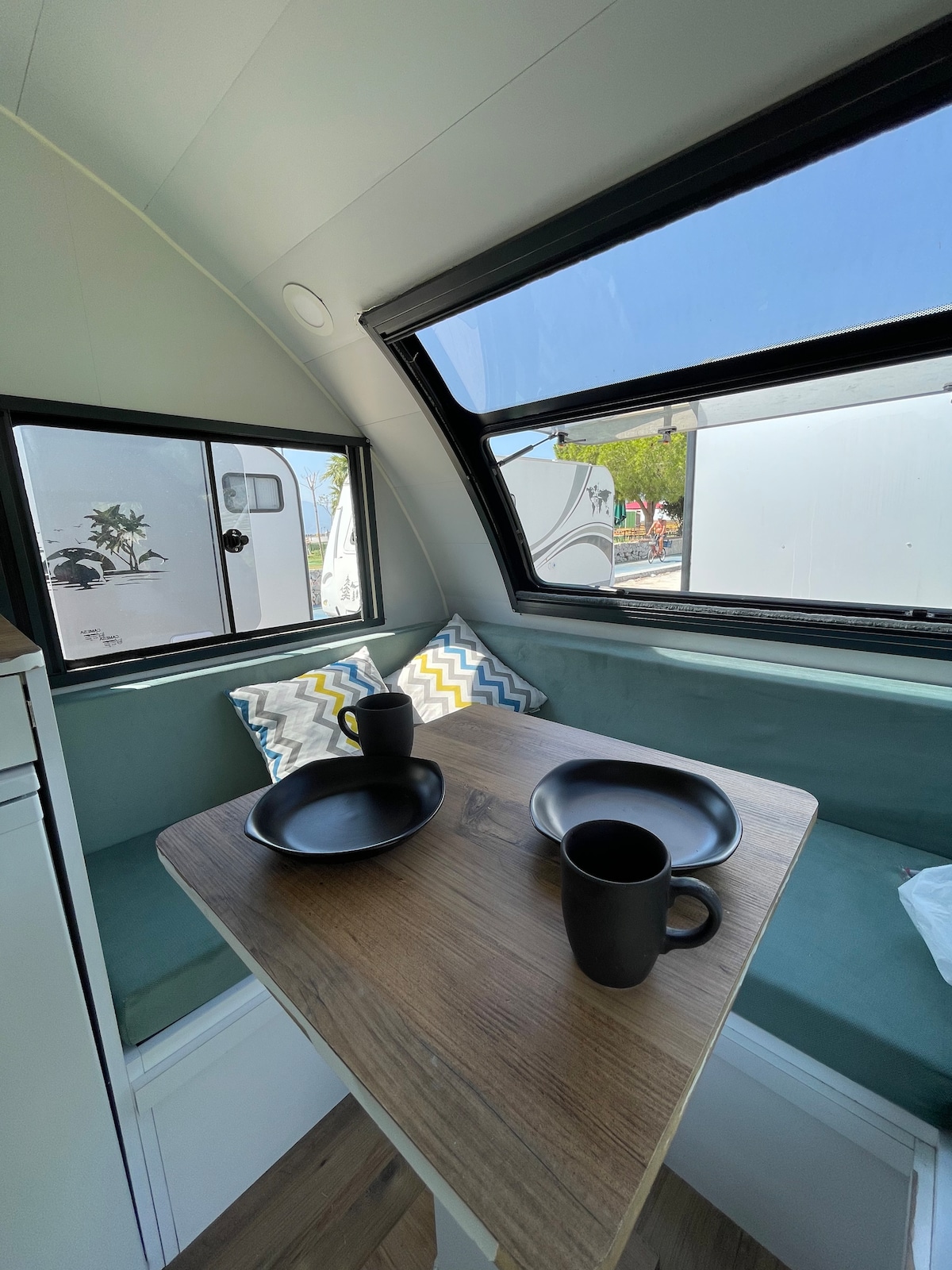 A bright dining area is featured, with large windows providing natural light. The space includes a wooden table topped with two black plates and mugs, accompanied by patterned cushions on the seating. Clean lines and a minimalist design are evident throughout.