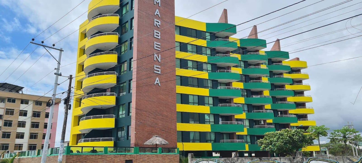 A modern multi-story building is presented, featuring bold yellow and green balconies. The structure showcases a prominent sign with the name of the building at the front. Surrounding greenery and a shaded area suggest a welcoming outdoor space for residents and visitors.
