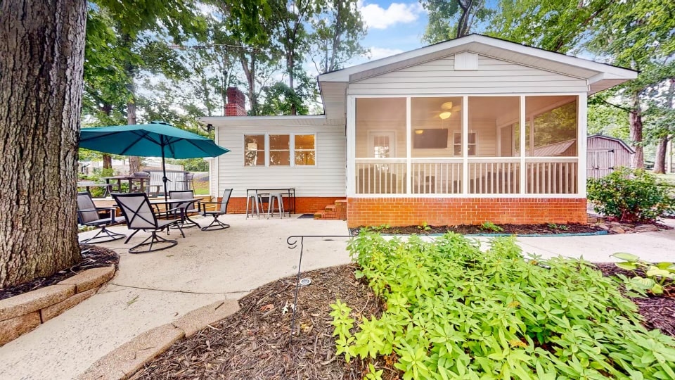 A screened porch is featured at the front of the house, surrounded by large windows for natural light. A patio area includes outdoor seating and an umbrella, alongside a pathway bordered by low shrubs and landscaping. Tall trees provide shade and a serene atmosphere.