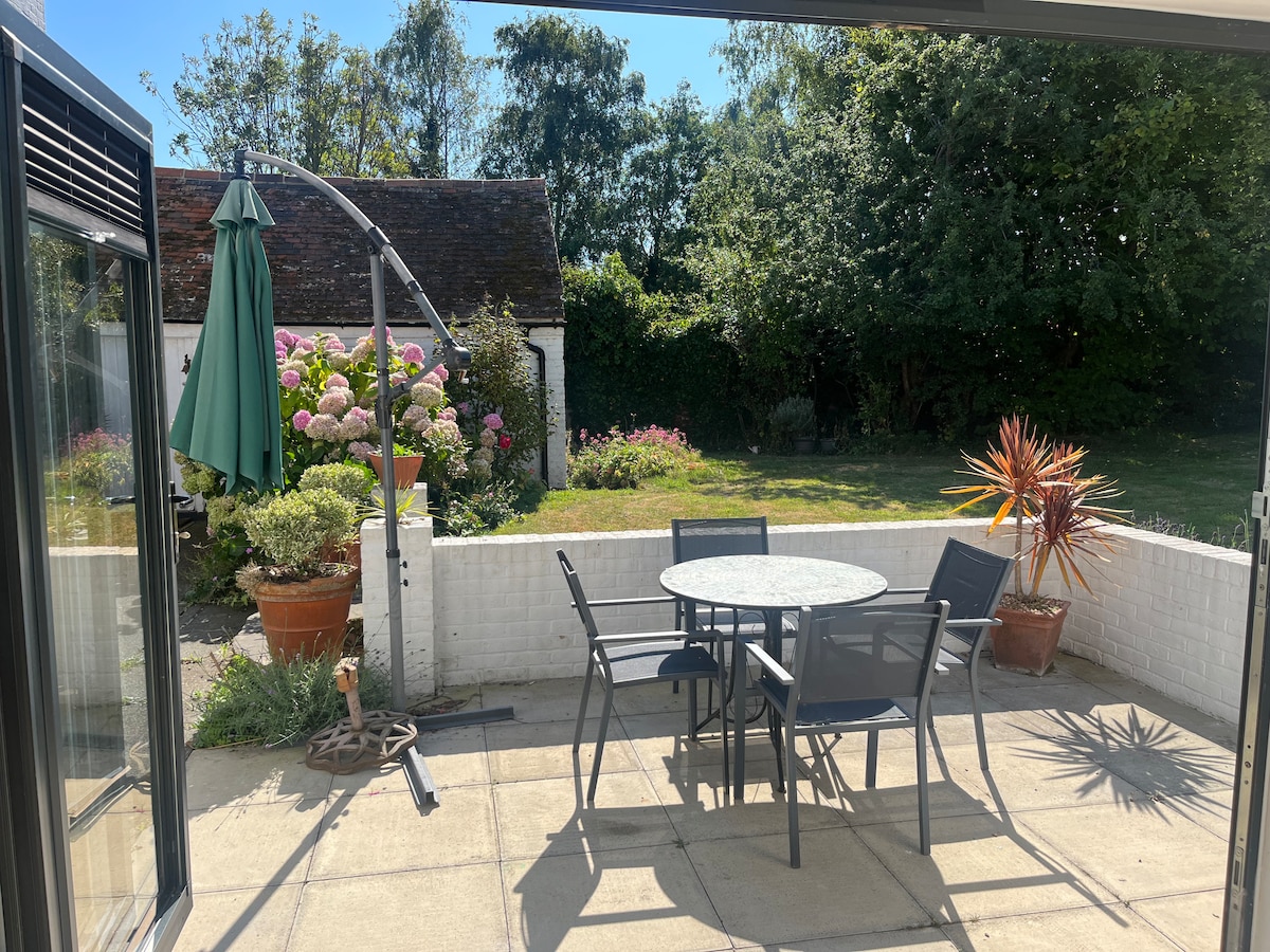 An outdoor patio area is shown, featuring a round table surrounded by four chairs. A green umbrella provides shade, while several potted plants enhance the space. In the background, a well-maintained garden with blooming flowers and lush greenery is visible.