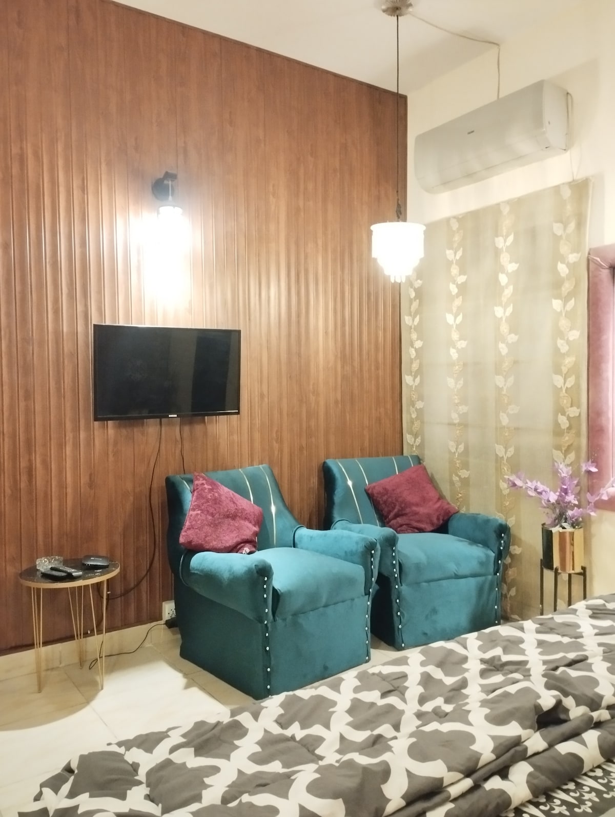 A cozy corner in the room features two upholstered chairs in a teal hue, with decorative cushions in rich burgundy. A wall-mounted flat-screen TV is positioned for viewing, while soft lighting accents the textured wooden wall behind, enhancing the inviting atmosphere.