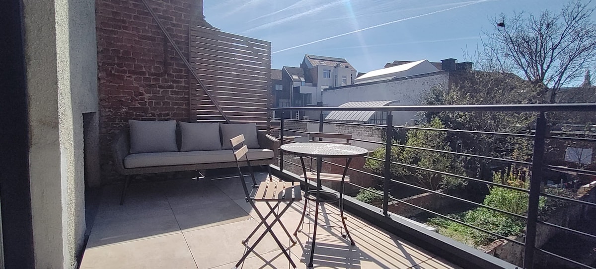 A private terrace is visible, featuring a comfortable gray sofa and a small round table accompanied by a folding chair. The surroundings showcase a mix of brick walls and greenery under a clear blue sky, allowing for a bright and open atmosphere.
