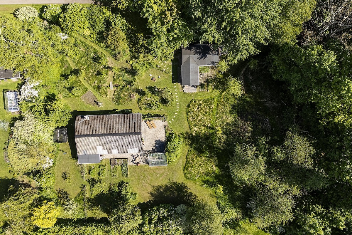 An aerial view captures a lush green landscape surrounding a rustic summer house. The expansive garden features walking paths, planting areas, and a greenhouse. Adjacent to the main structure is a smaller building, both situated among trees and natural vegetation, inviting tranquility and outdoor exploration.
