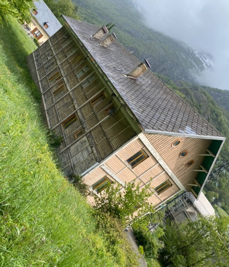 A renovated chalet is nestled among lush greenery, displaying a combination of wooden textures and natural surroundings. The exterior features large windows and a sloped roof, creating a harmonious blend with the mountainous landscape. Nearby, misty hills are visible in the background.