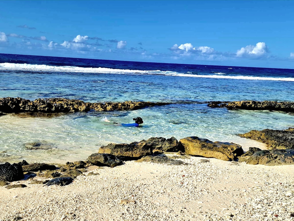 A serene beach scene is captured, featuring clear blue waters gently lapping against the sandy shore. Rocky formations create natural borders, with one individual enjoying the shallow water. The sky is bright with scattered clouds, enhancing the tranquil atmosphere of the coastline.
