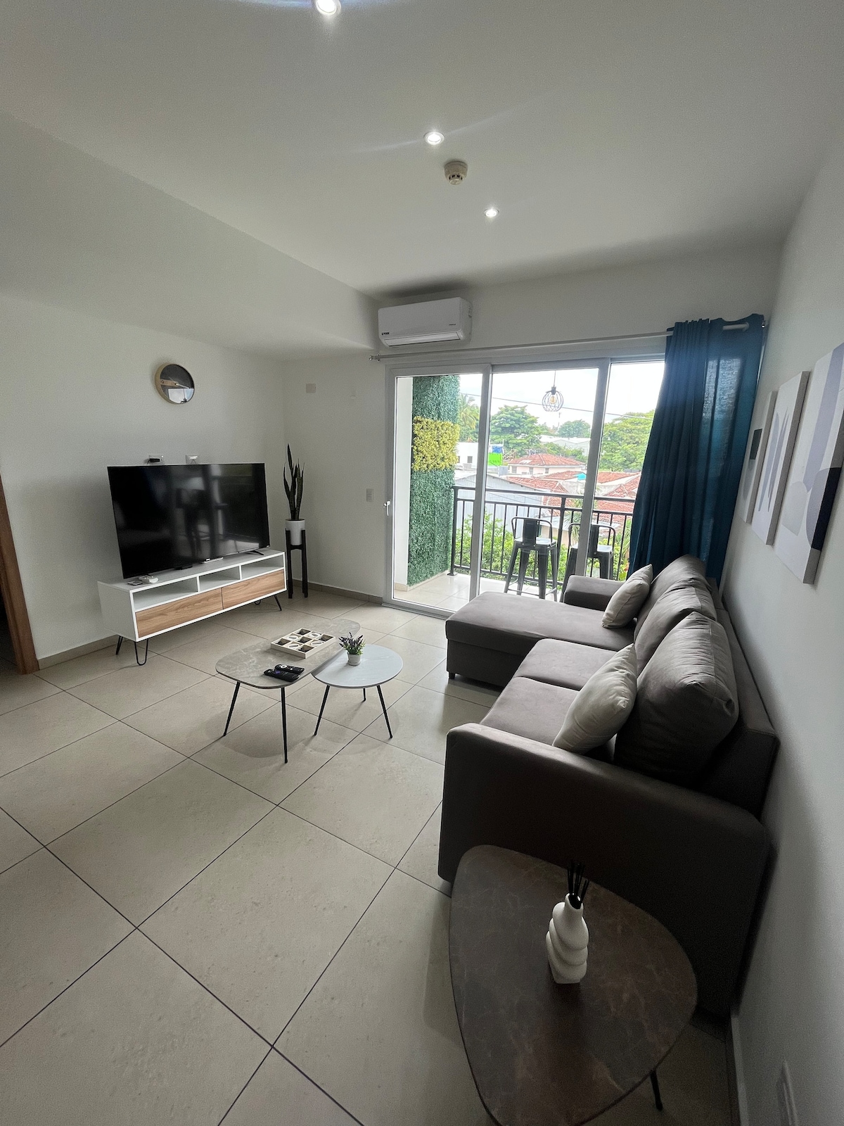 A modern living area is furnished with a comfortable gray sofa and two coffee tables. A flat-screen TV is positioned across from the seating. Natural light flows through large glass doors that open to a small balcony, creating a bright and inviting space.