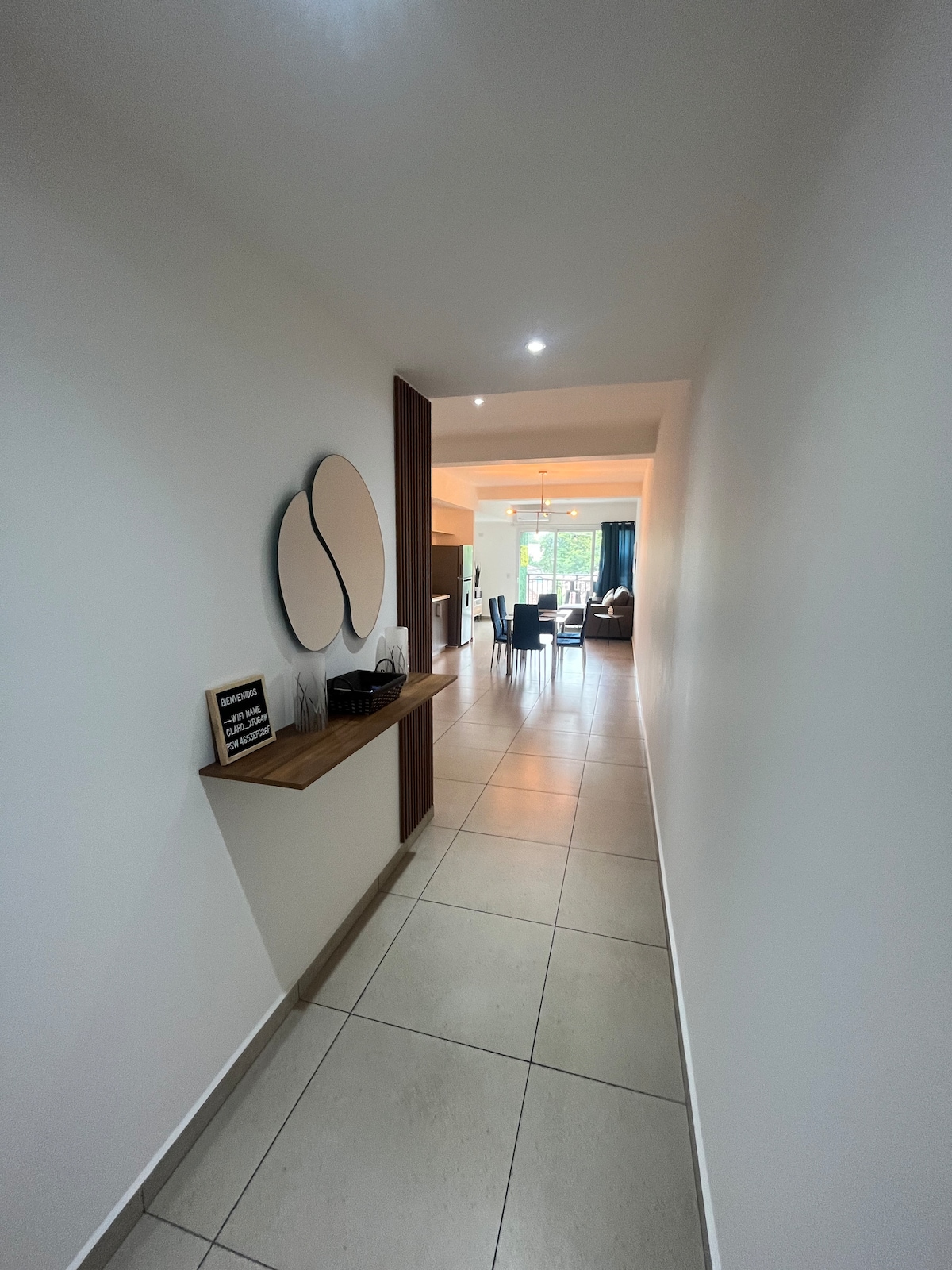 A modern hallway offers a glimpse into a spacious, well-lit living area at the end. Neutral tones and sleek tile flooring lead to a dining and sitting area, while a decorative wall piece adds a subtle touch of elegance.