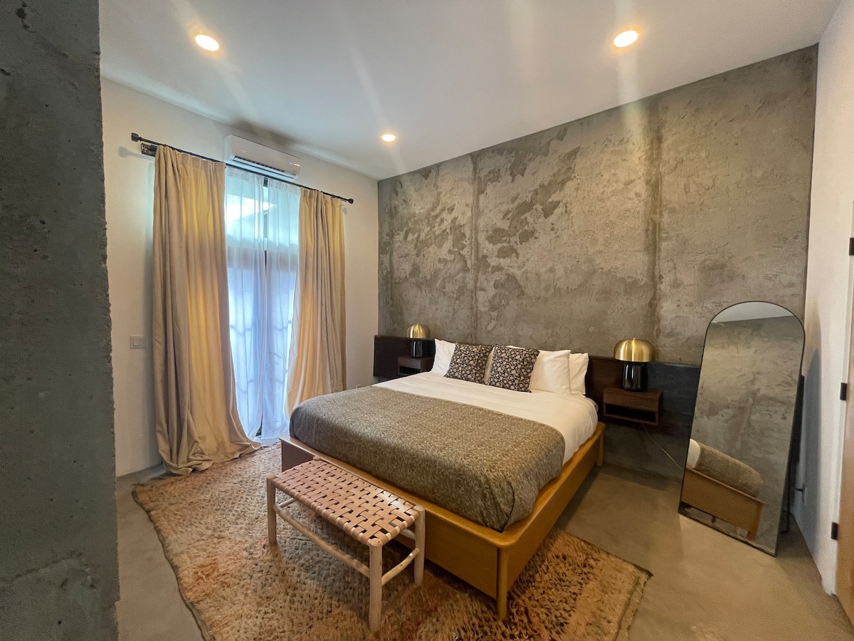 The king bedroom features textured concrete walls and a large window covered by sheer curtains. A spacious bed with neutral bedding is complemented by two bedside tables with lamps. A decorative mirror and a woven rug add warmth to the minimalist design.