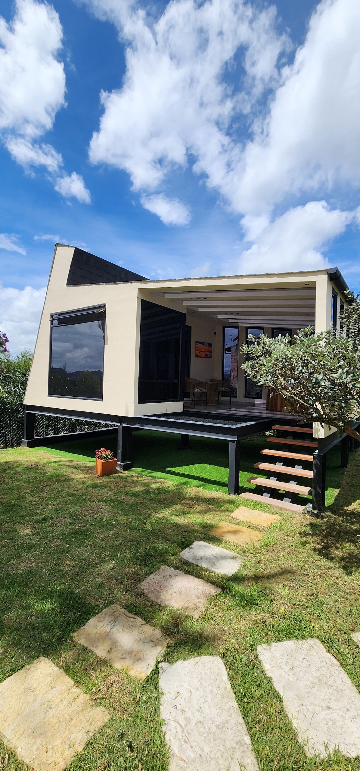 A modern glamping structure features a distinctive angled roof and large windows, allowing natural light to fill the interior. A set of wooden steps leads to the entrance, while a lush green lawn surrounds the home, complemented by stone pathway tiles.