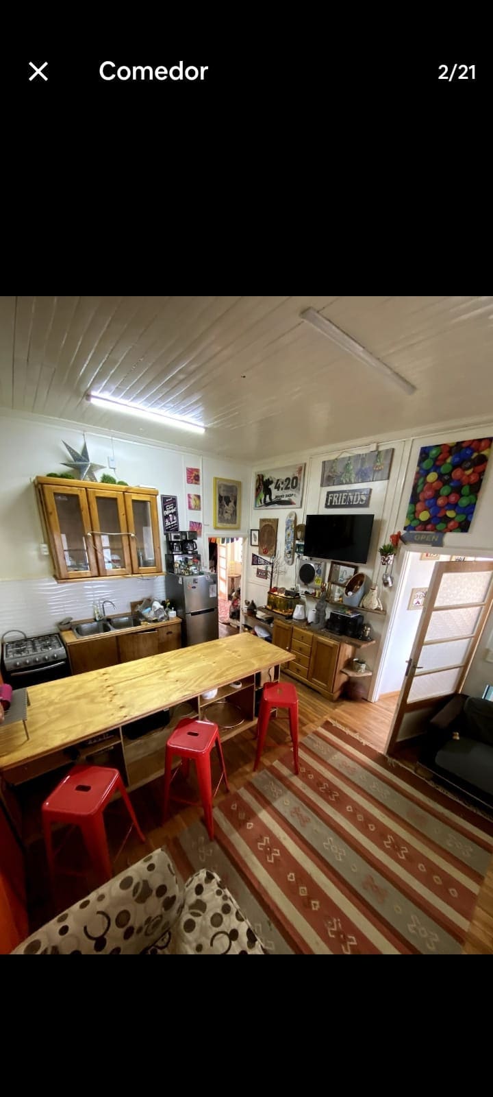 A bright kitchen area is visible, featuring wooden cabinetry and a spacious table surrounded by red stools. Art adorns the walls, adding character. The area is well-lit with natural light, highlighting various kitchen essentials and decorative elements.