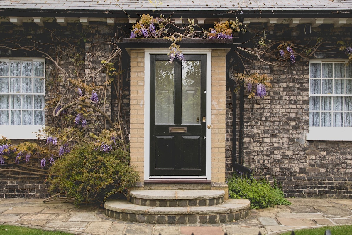 A charming entrance features a dark front door framed by light bricks and surrounded by blooming wisteria vines. Two windows with white curtains flank the doorway, while a small garden area with greenery adds to the inviting look of the entryway.