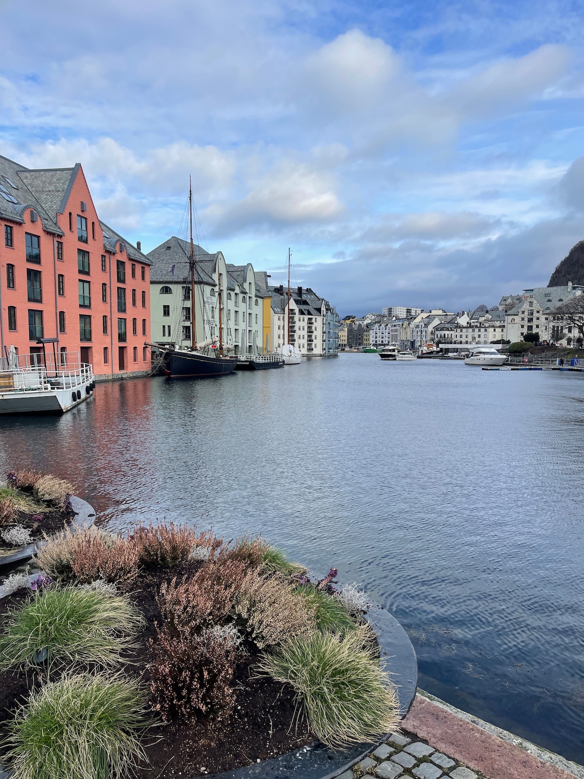 A serene waterfront scene displays colorful buildings lining the river. Soft clouds hover above, creating a tranquil atmosphere. Boats are gently docked along the shore, while lush gardens are visible in the foreground, adding natural elements to the urban landscape.