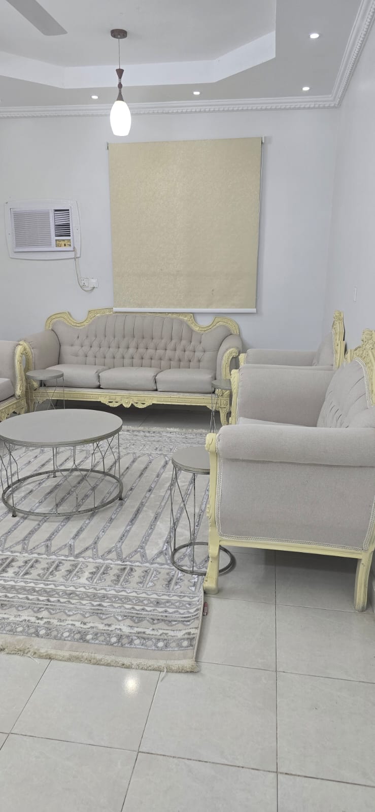 A stylish living area features elegant seating arranged around a low coffee table. The couch and chairs are upholstered in soft beige fabric with ornate yellow accents. A light rug with subtle patterns lies underneath, complementing the room's bright ambiance.