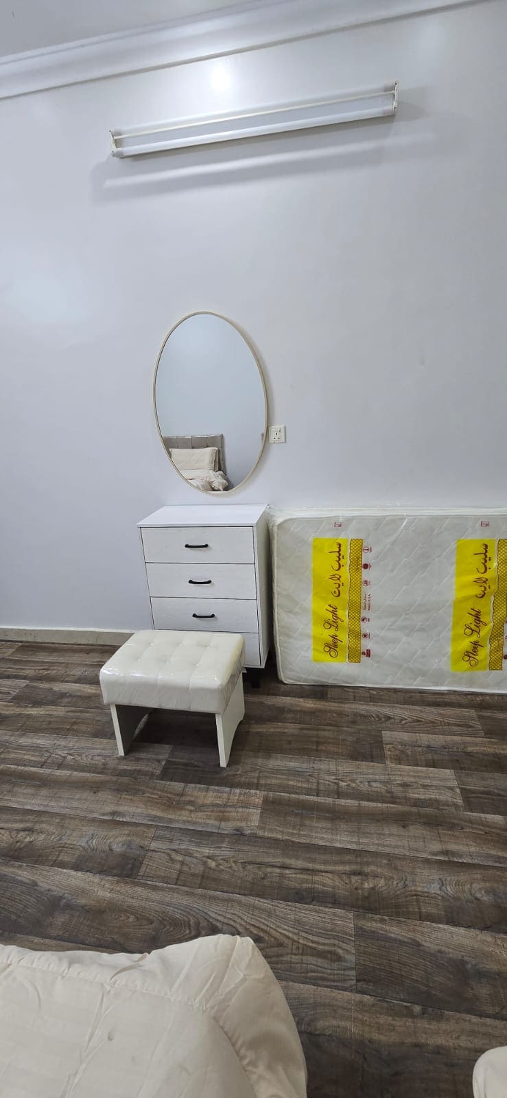 A simple bedroom space features a light-colored dresser with four drawers and a circular mirror above it. A small, upholstered stool is positioned nearby. A new mattress wrapped in protective covering leans against the wall, complementing the wooden floor.