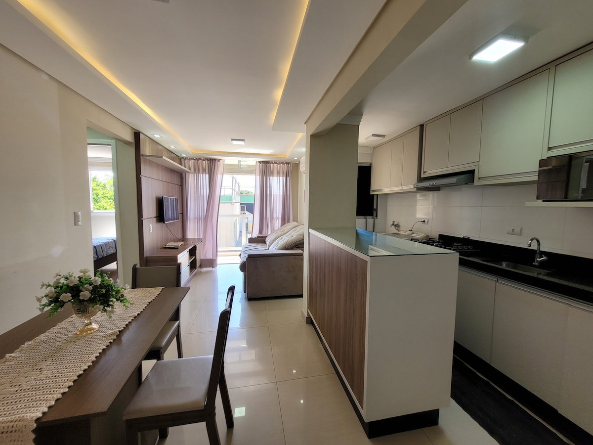 An open-plan living space is presented, featuring a modern kitchen with cabinetry and appliances. A dining table with chairs occupies one side, while a comfortable sofa faces a wall-mounted TV. Natural light filters through large windows adorned with sheer curtains.