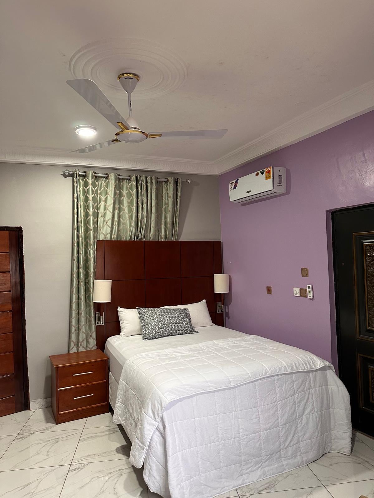 A cozy bedroom is highlighted by a spacious bed with crisp white bedding. A matching bedside table sits beside the bed, and two lamps provide soft lighting. The walls are painted in a soothing purple, complemented by patterned curtains that frame the window.