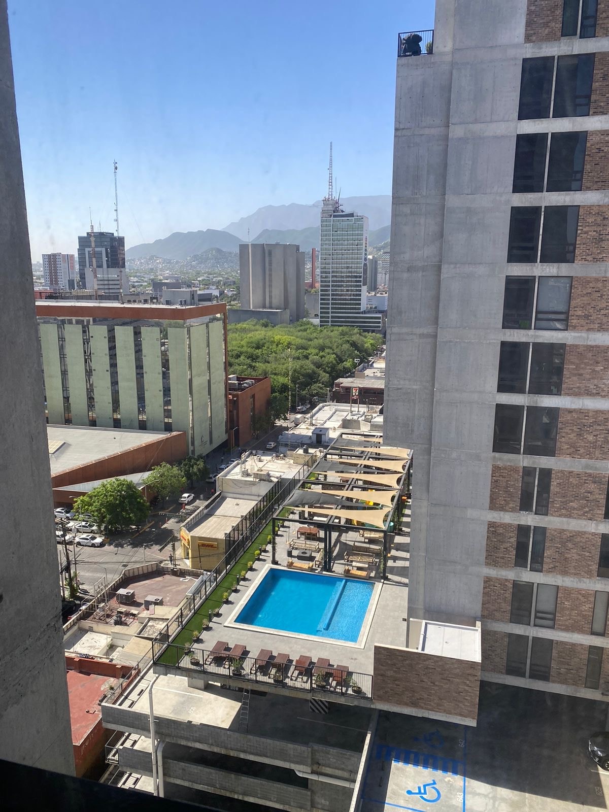 An aerial view of a vibrant city landscape is displayed, featuring a clear blue pool surrounded by lounge chairs. Towering buildings and distant mountains contribute to the urban scenery, with streets lined by greenery below. Sunlight enhances the inviting outdoor space.