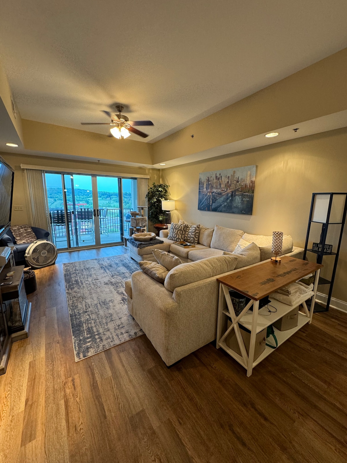 A spacious living area showcases a comfortable cream-colored sofa and a wood accent table. A large window offers a view of the outdoors, allowing natural light to brighten the space. A television is placed on a stand, complementing the overall design.