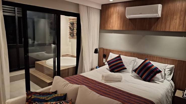 A modern bedroom features a spacious bed adorned with colorful accent pillows. A comfortable seating area is visible in the foreground. Floor-to-ceiling glass doors lead to a private bathroom area, which includes a sleek, round soaking tub, further enhancing the room's contemporary design.