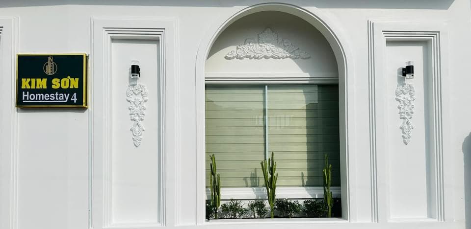 The exterior facade features a white color scheme, accented with decorative elements flanking a large window. Cacti are visible in the planter below, creating a fresh and inviting appearance. A sign with the text 'KIM SƠN Homestay 4' is prominently displayed to the left.