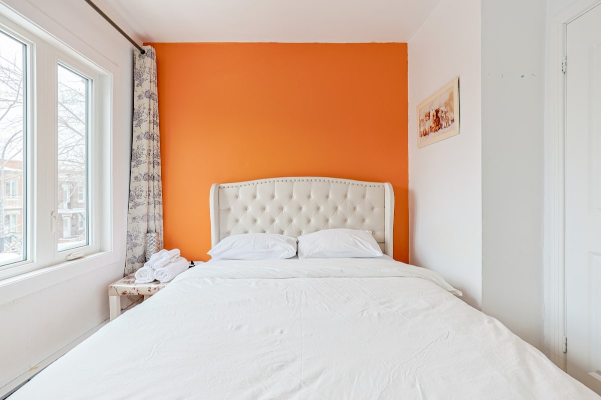 A spacious bed with a tufted headboard is set against an orange wall, providing a vibrant contrast. Natural light enters through two large windows, while a small bedside table is positioned nearby. Soft linens cover the bed for a serene look.