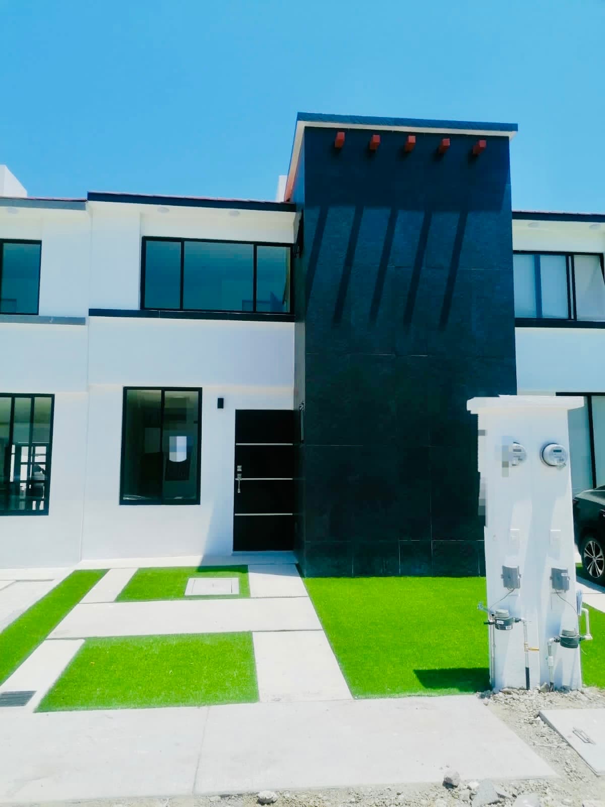 A modern two-story building showcases a sleek black facade contrasted by white walls. The green grass in the foreground adds a touch of color. Large windows line the front, inviting natural light into the space. Utilities are neatly provided on one side.