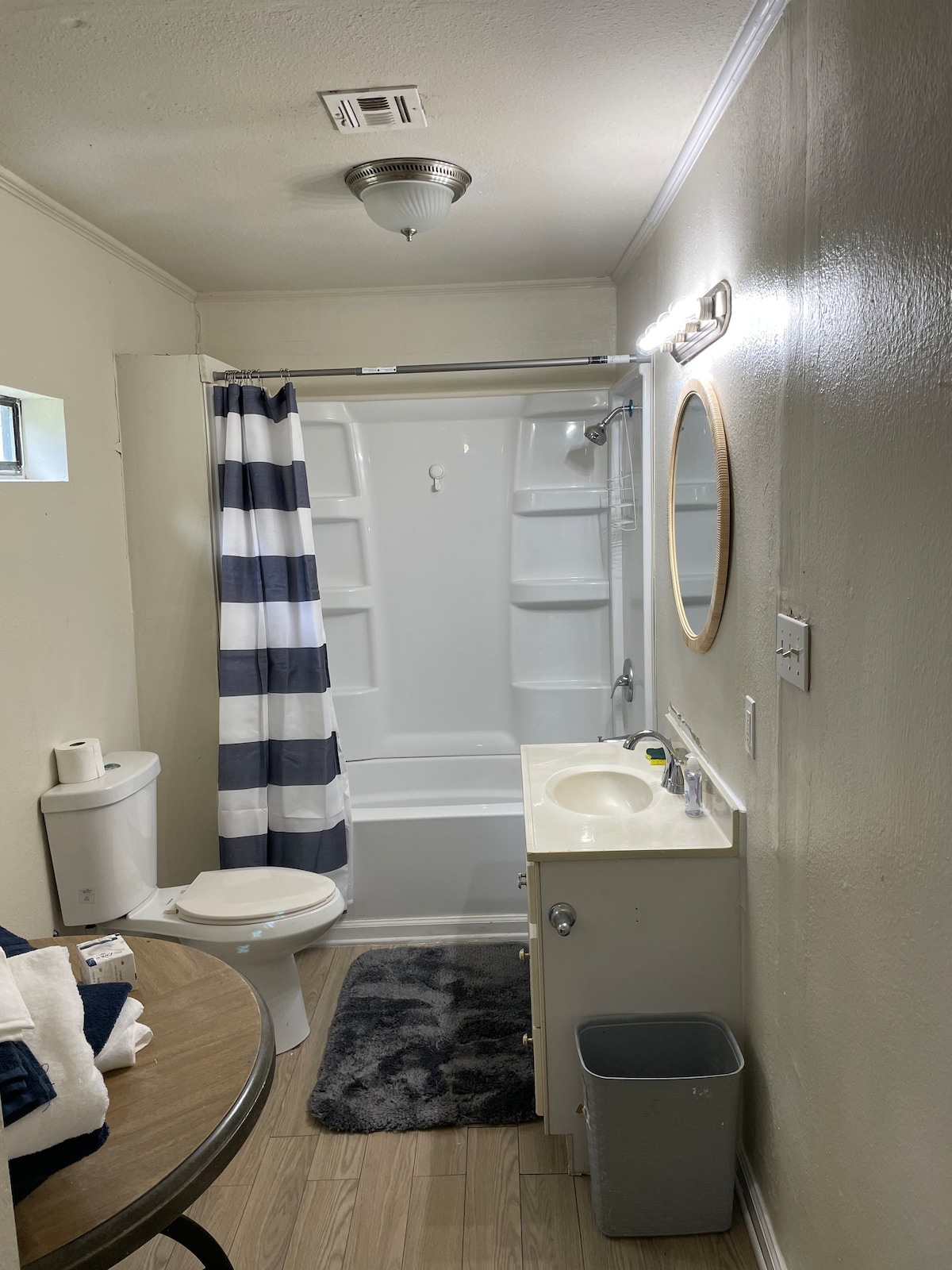 The bathroom features a clean design with a shower equipped with a white curtain having navy blue stripes. A round mirror is mounted above a light-colored vanity, which holds basic amenities. A small rug lies on the floor beside a toilet and waste basket.