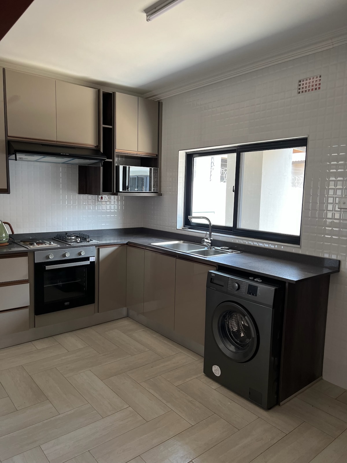 A modern kitchen features sleek cabinetry with a mix of light and dark finishes. Stainless steel appliances including a stove and dishwasher are present. A sink is positioned beneath a window, allowing natural light to enter, while a washing machine is discreetly located nearby.