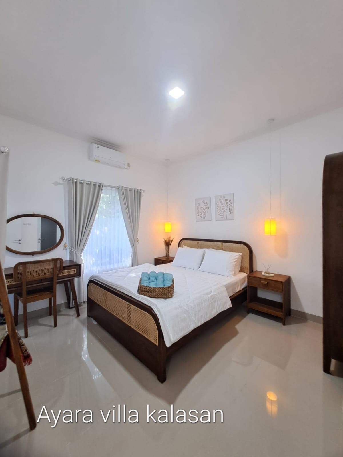 A well-lit bedroom features a spacious bed with a white quilt. Soft drapes frame a window allowing natural light. A wooden desk and mirror are positioned beside the bed. Decorative wall art adds a touch of character, while two pendant lights provide gentle illumination.