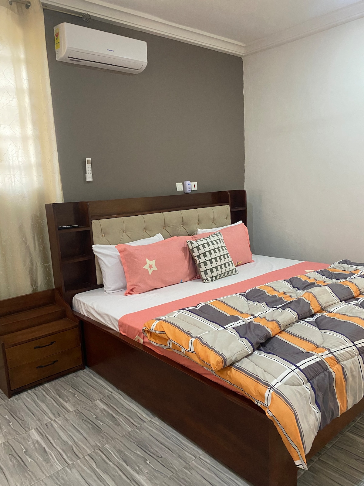 A cozy sleeping area is equipped with a spacious bed featuring a headboard and built-in side shelves. Light-colored bedding contrasts with a patterned blanket. An air conditioning unit is mounted on the wall, and soft natural light enters through a nearby window dressed with sheer curtains.