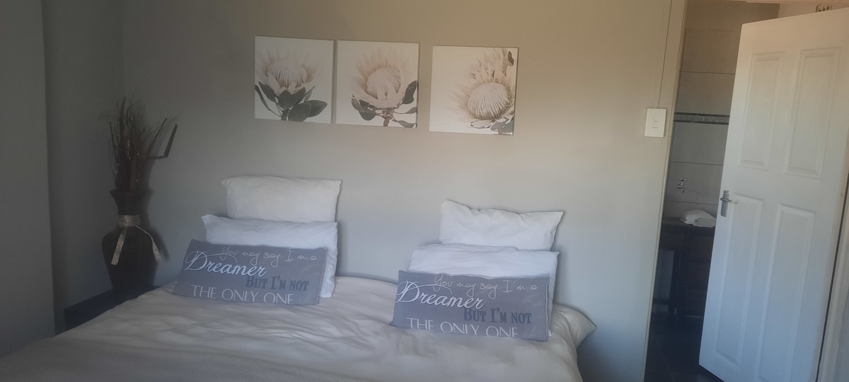 A serene bedroom layout features two spacious beds topped with white pillows and decorative throw pillows. On the wall, three floral art prints add a touch of nature. A subtle vase with dried plants stands in the corner, complementing the light grey walls.