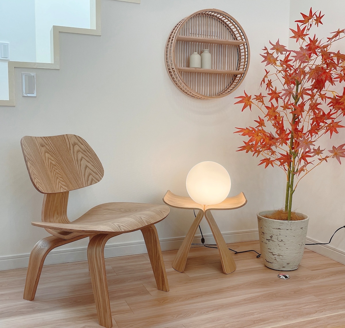 A cozy corner features a wooden chair beside a small table with a round lamp. An orange maple tree adds a touch of color, while decorative items are displayed on a circular wall shelf nearby. The light wood flooring enhances the bright atmosphere.