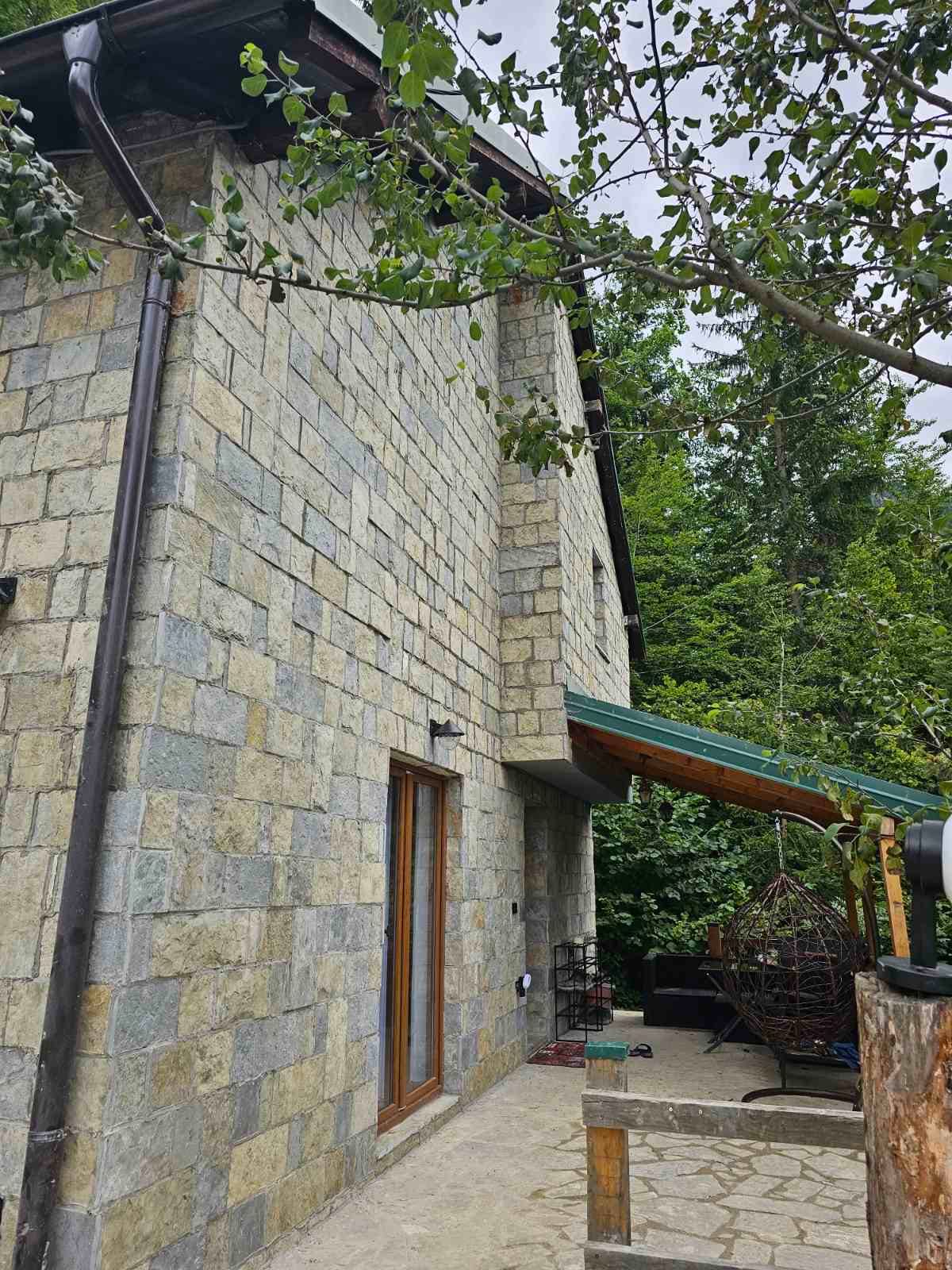 The exterior of a stone-clad house is surrounded by lush greenery, showcasing a combination of natural textures. A covered porch with a green roof leads to a cozy seating area, partially shaded by trees and featuring a hanging chair.