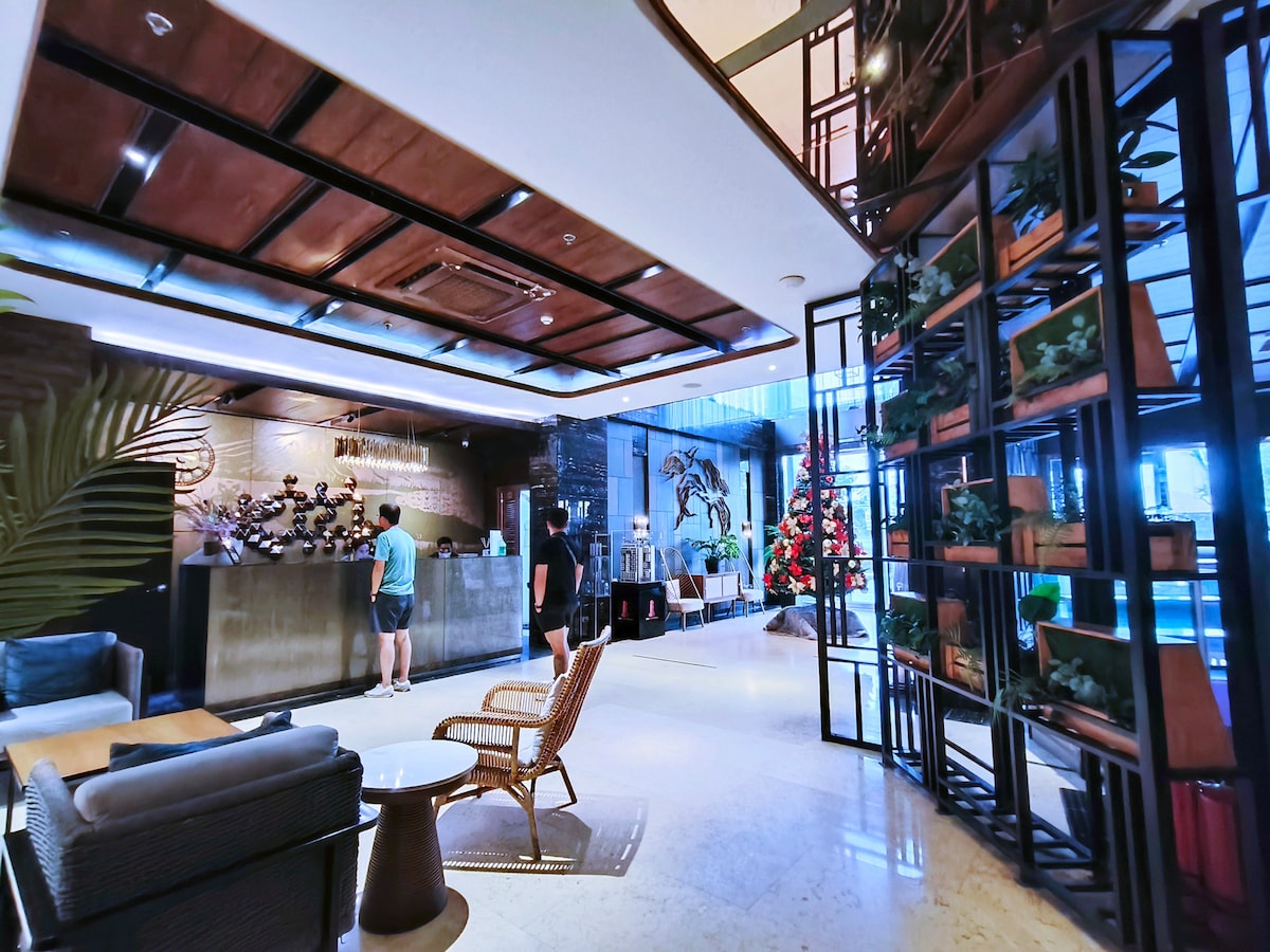 The lobby area features a reception desk with a modern design, complemented by decorative plants and art pieces. Comfortable seating options are arranged near a table, while large windows allow natural light to illuminate the space. A holiday tree adds a festive touch.