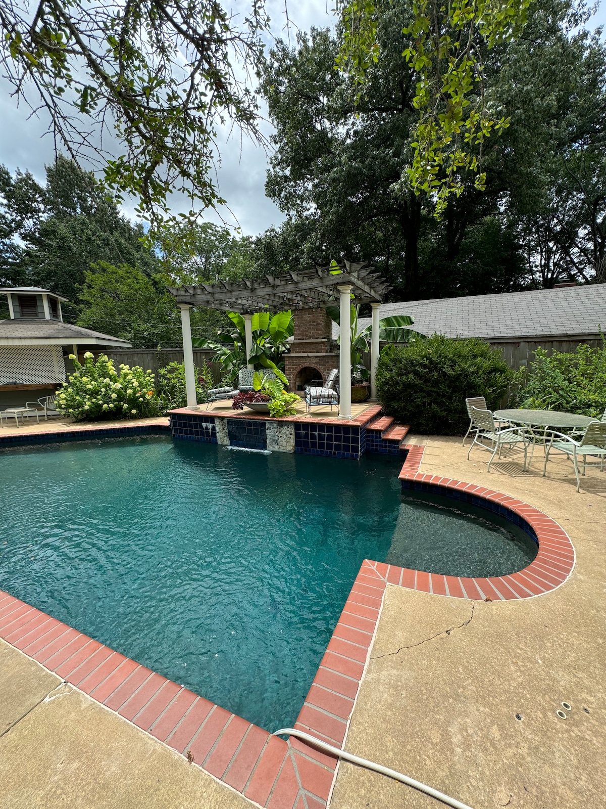 A private pool area is showcased, featuring a spacious swimming pool with a brick border. A raised deck with patio seating overlooks the water. Lush greenery surrounds the area, enhancing the tranquil setting, with a rustic stone fireplace providing a focal point.
