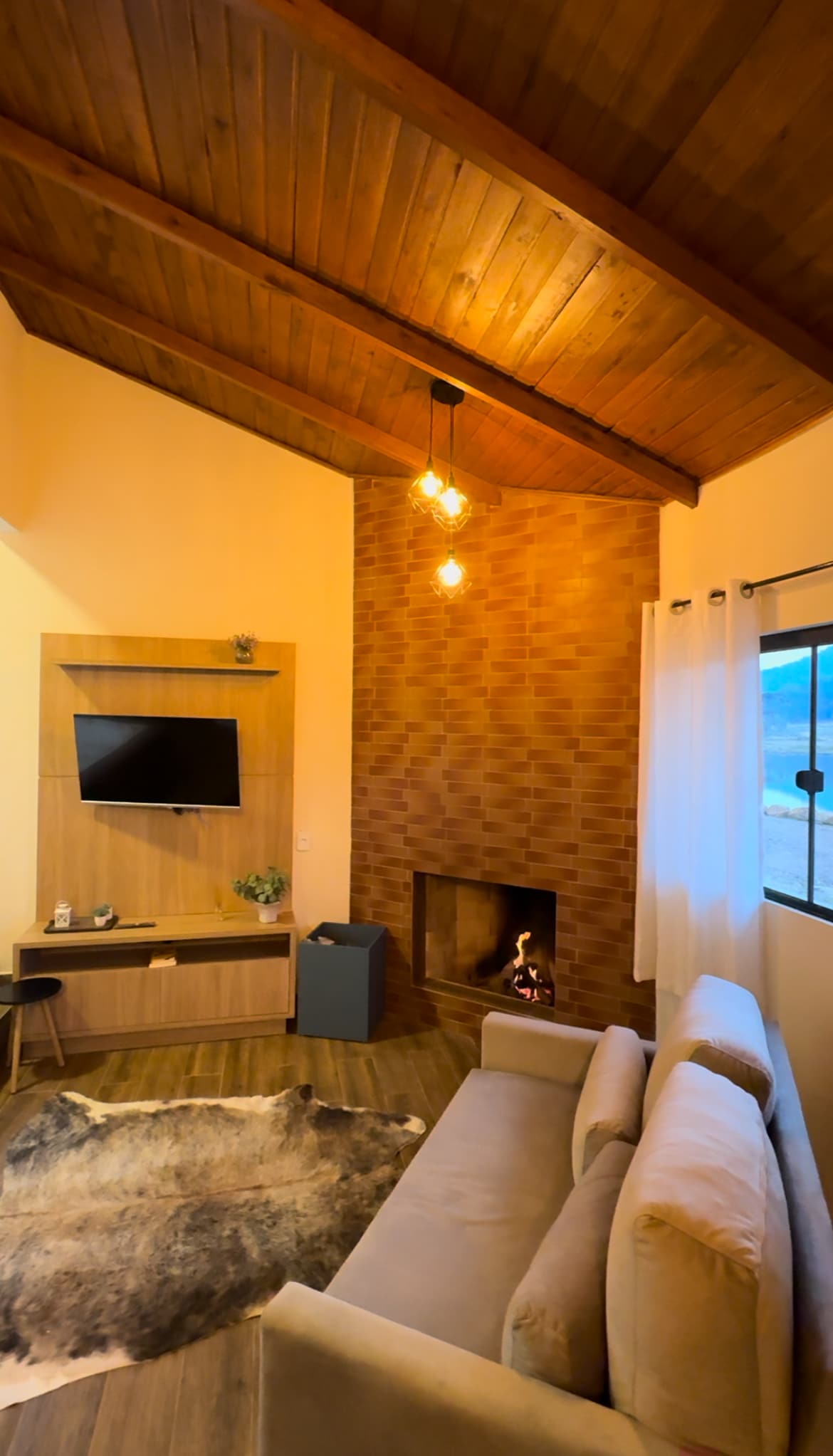 The living area features a beige sofa positioned next to a wood-burning fireplace, which is set within a brick wall. A television is mounted above a wooden console that includes decorative plants. Natural light filters through a nearby window, enhancing the warm ambiance.