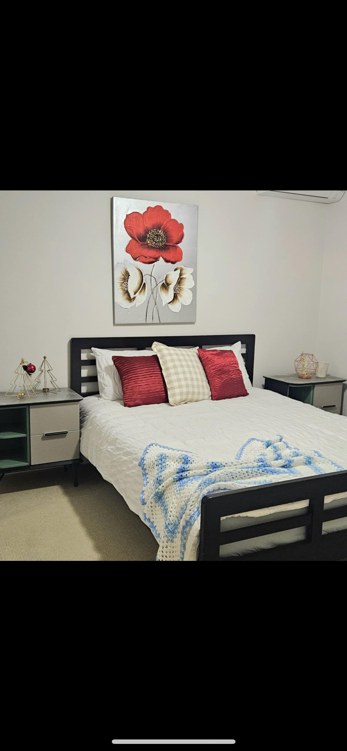 A well-appointed bedroom features a spacious bed dressed with white linens and decorative throw pillows in red and tan. A floral wall art piece adds color above the bed. Nightstands with lamps are positioned on either side, complementing the contemporary design.
